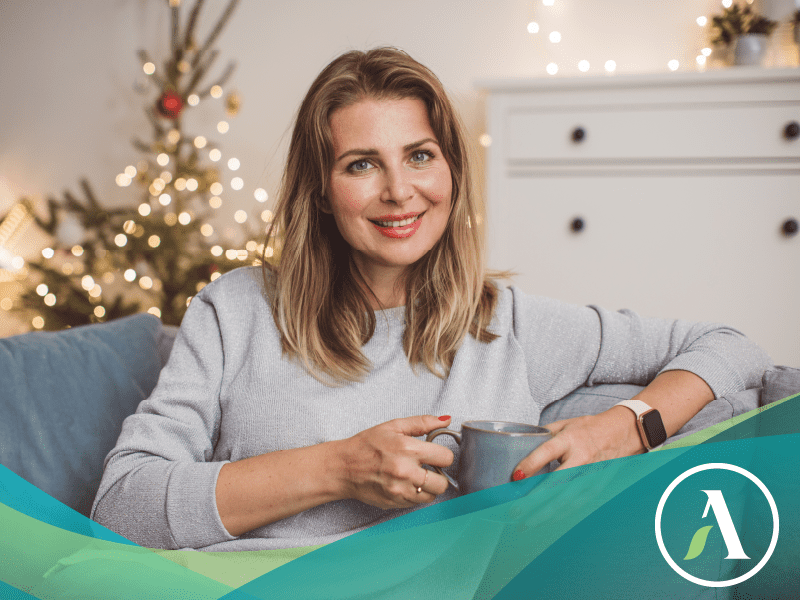Caucasian woman smiles at camera holding a mug while sitting on a couch. Christmas decorations behind her.