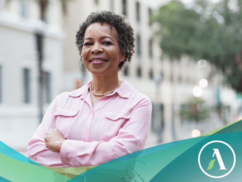 Black woman smiles at camera with arms crossed, standing outside in an urban setting.